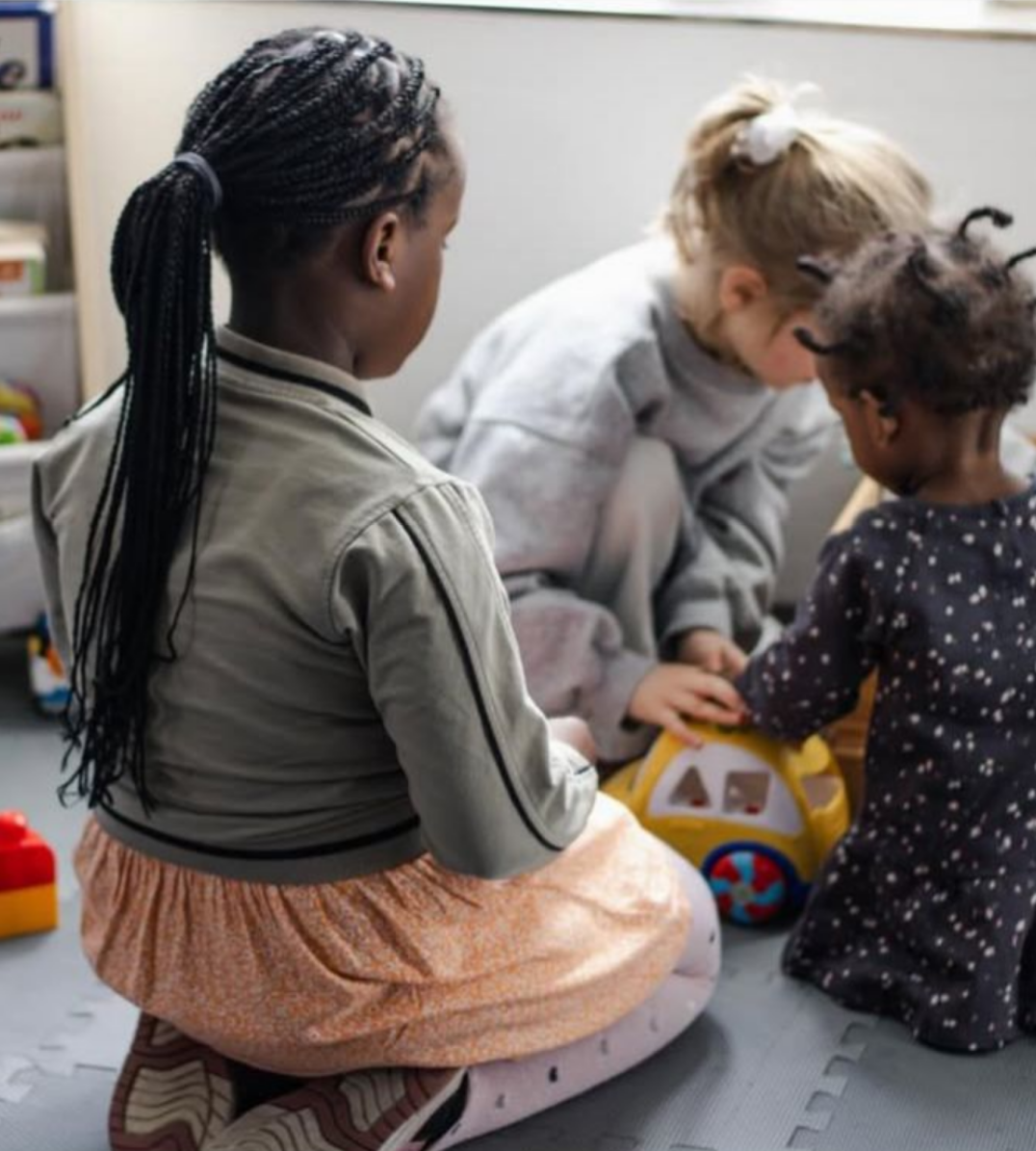 Drei kleine Kinder sitzen auf dem Boden und spielen gemeinsam mit einem bunten Spielzeugauto; sie sind einander zugewandt und konzentriert, in einer ruhigen, freundlichen Umgebung.