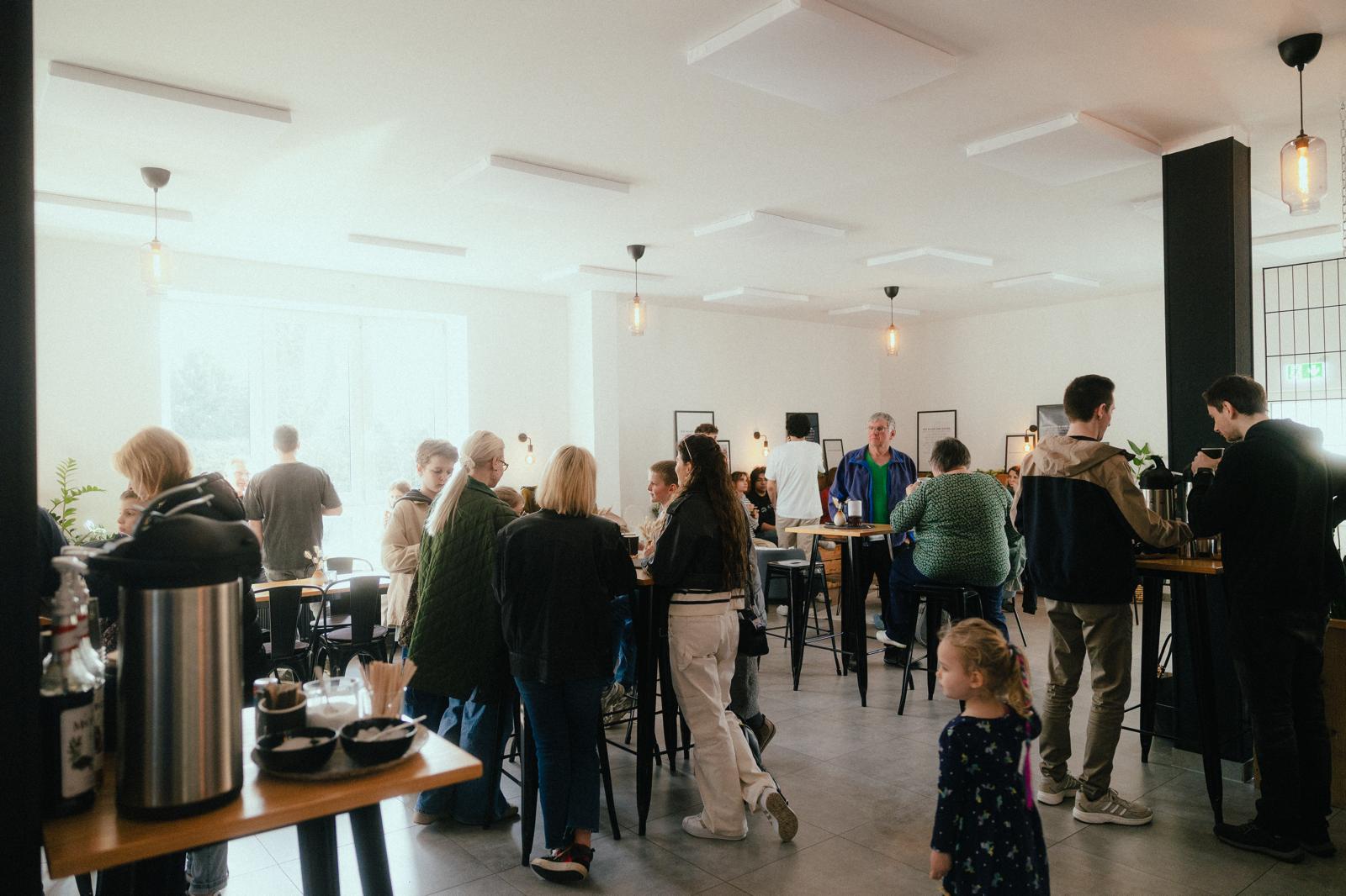 Helles, modernes Café im Riverside Church: Viele Menschen stehen und sitzen an hohen Tischen, unterhalten sich und trinken Kaffee; im Vordergrund steht ein kleines Kind, im Hintergrund fällt viel Tageslicht durch große Fenster.